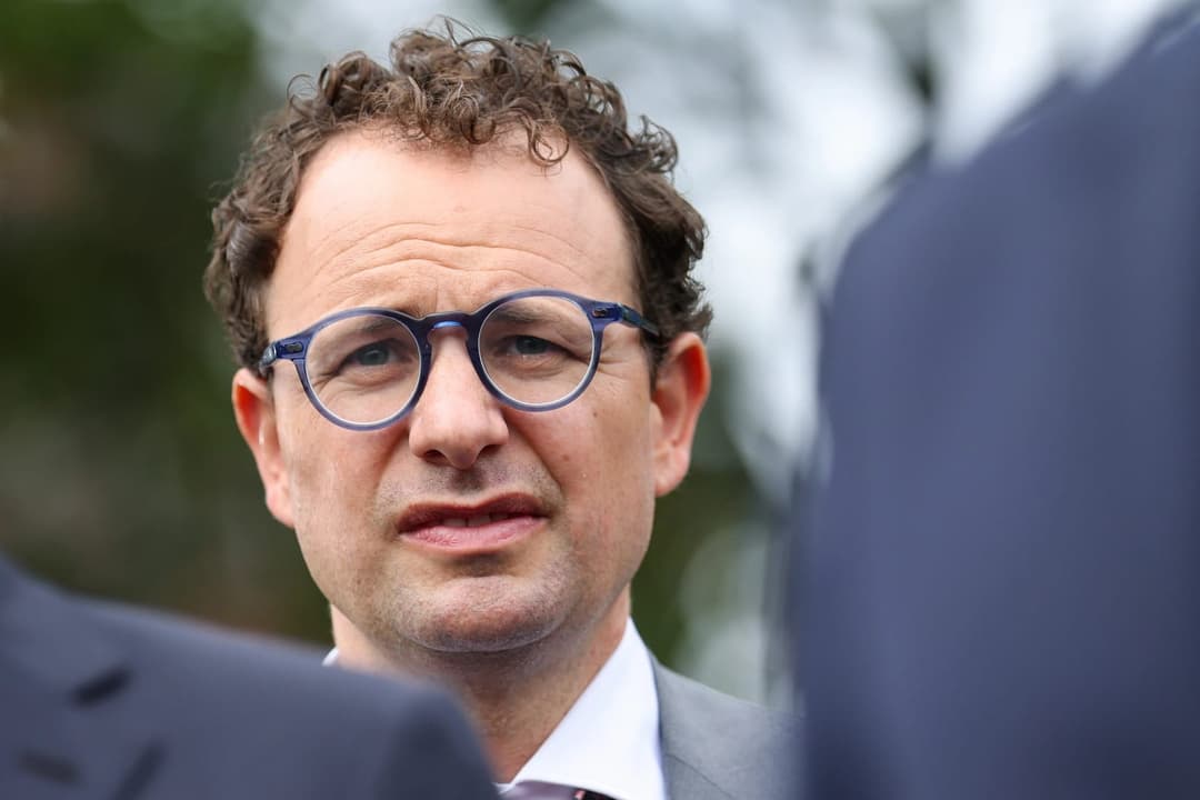 The image shows a man with curly brown hair and glasses, wearing a gray suit jacket and a white collared shirt. He is standing in front of a blurred background, which appears to be a natural setting with trees and foliage. The man's facial expression suggests he is speaking or reacting to something, and his gaze is dir