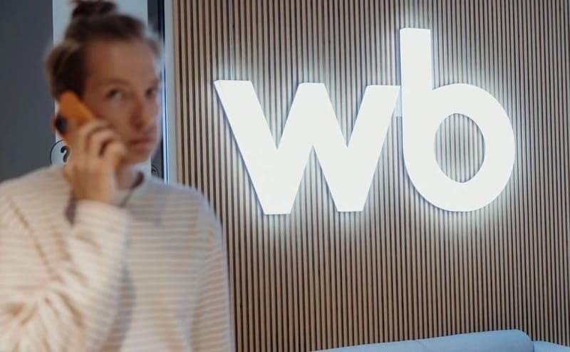 The image shows a person standing in front of a wall with vertical wood paneling. The person is wearing a light beige or cream-colored striped shirt and is holding a phone to their ear. The wall behind them features a large, illuminated "wb" logo. The person's hair is pulled up in a bun. The image is taken from a sligh