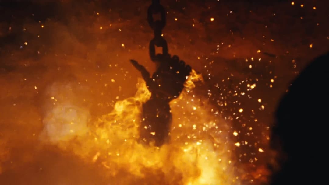 The image shows a dark silhouette of a hand gripping a chain, suspended in the air against a backdrop of intense orange and yellow flames. The hand and chain are the central focus of the image, with the flames providing a dramatic contrast in color and intensity. The background is filled with small, bright sparks, crea
