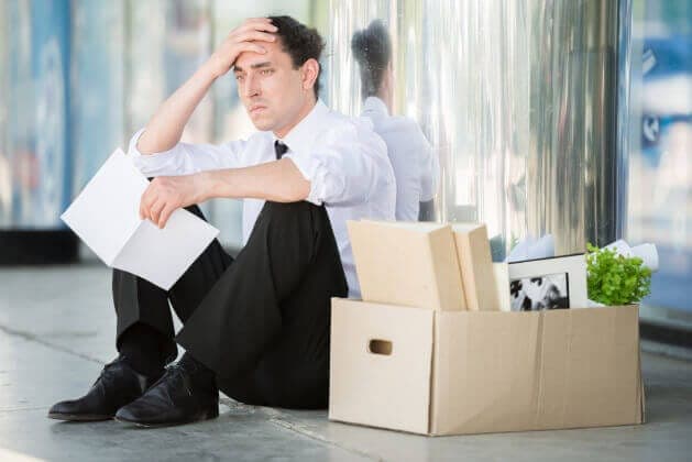 The image shows a man sitting on the ground in front of a building with a glass facade. He is dressed in a white shirt and black pants, and is holding a piece of paper in his left hand. His right hand is resting on his head, suggesting a state of distress or frustration. Next to him is a cardboard box containing variou
