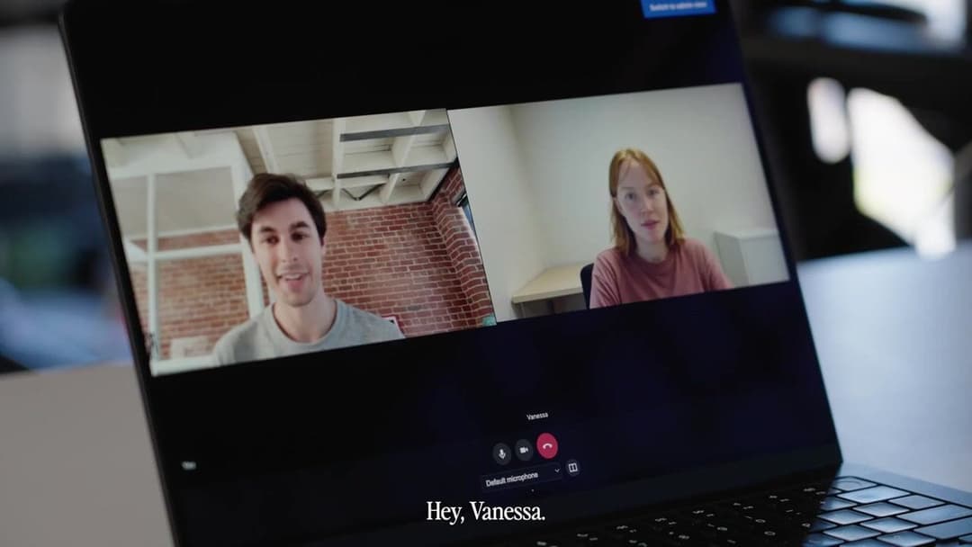 The image shows a laptop displaying a video call interface. The screen shows two individuals, a man and a woman, engaged in a video call. The man is wearing a gray t-shirt and has short brown hair. The woman is wearing a pink t-shirt and has long, light brown hair. The laptop's screen is dark, and the video call interf
