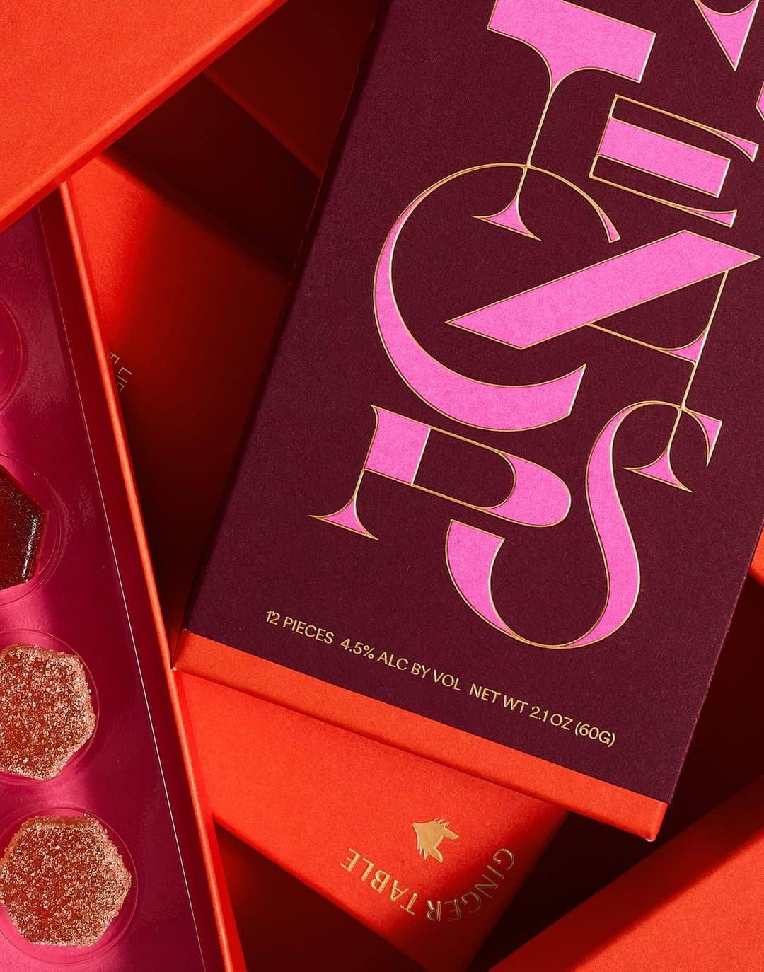 The image shows a collection of boxes, primarily in shades of red and orange, with a dark maroon or burgundy background. The boxes are stacked in a somewhat haphazard manner, creating a sense of depth and dimension. The topmost box is adorned with a pink and gold design that reads "GINZER TABLE" and "PIZZES", suggestin