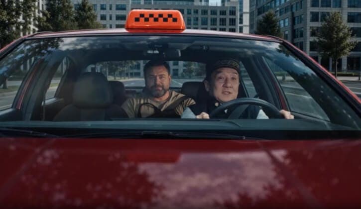The image shows two men seated in a red car, which appears to be a taxi, on a city street. The car's roof is adorned with a black and orange checkered taxi sign. The man driving the car is wearing a dark-colored cap and a dark-colored jacket. The man sitting in the passenger seat is wearing a light brown or tan shirt a