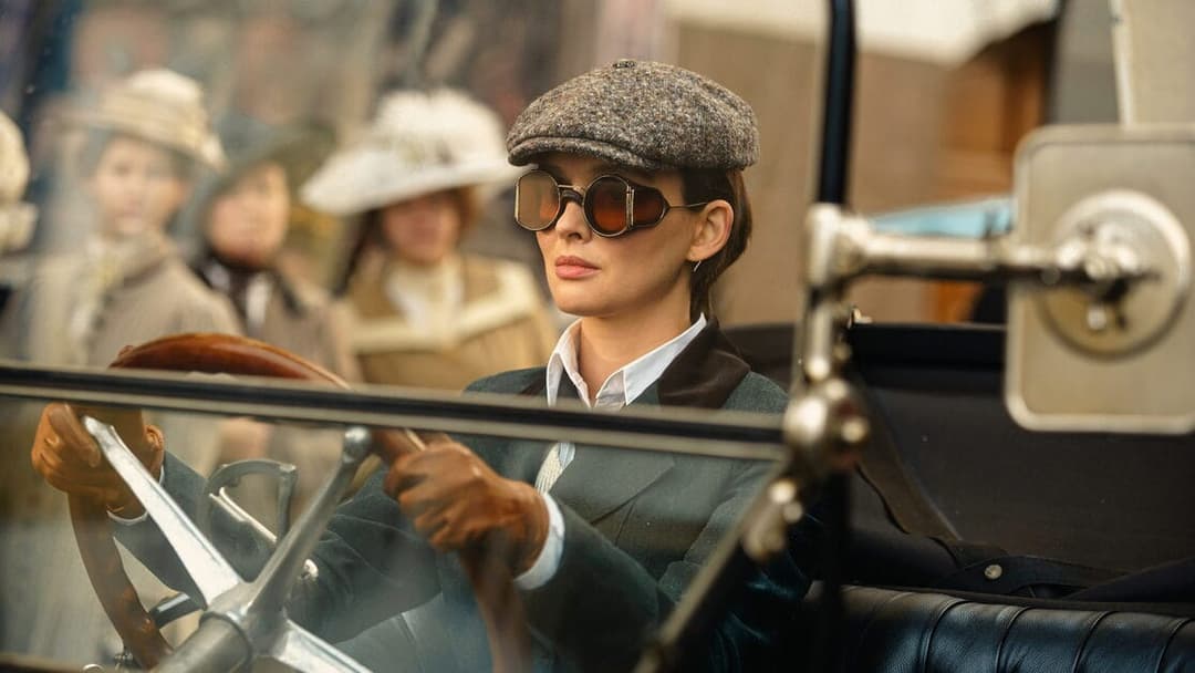 The image shows a woman seated in the driver's seat of a vintage car, possibly a Model T. She is wearing a dark green or teal jacket, a white collared shirt, and a gray tweed cap. She is also wearing brown sunglasses and brown gloves. The car's interior is dark, and the background is out of focus, suggesting a busy str