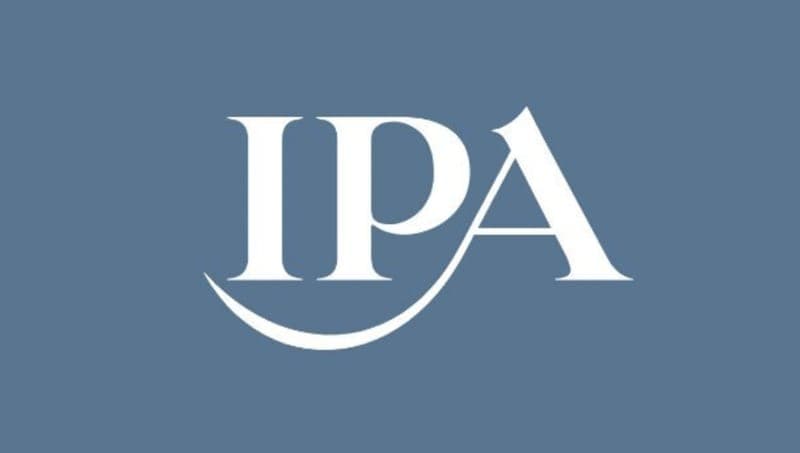 The image presents the logo of the International Press Association (IPA). The logo is a stylized representation of the letters "IPA" in white, set against a solid blue background. The letters are arranged in a way that gives the impression of a curved line or a smile, with the "A" slightly overlapping the "P" and the "