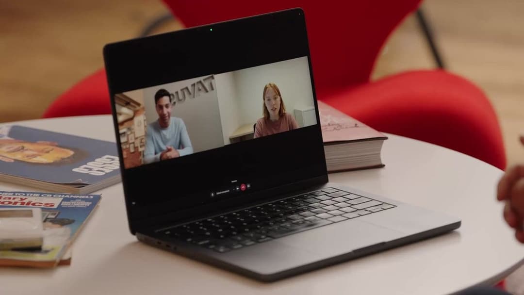 The image shows a laptop computer placed on a white table. The laptop screen displays a video call with two individuals, one male and one female, who appear to be engaged in a conversation. The male is wearing a light blue/grey t-shirt, and the female is wearing a pink/red shirt. The laptop is positioned in the center