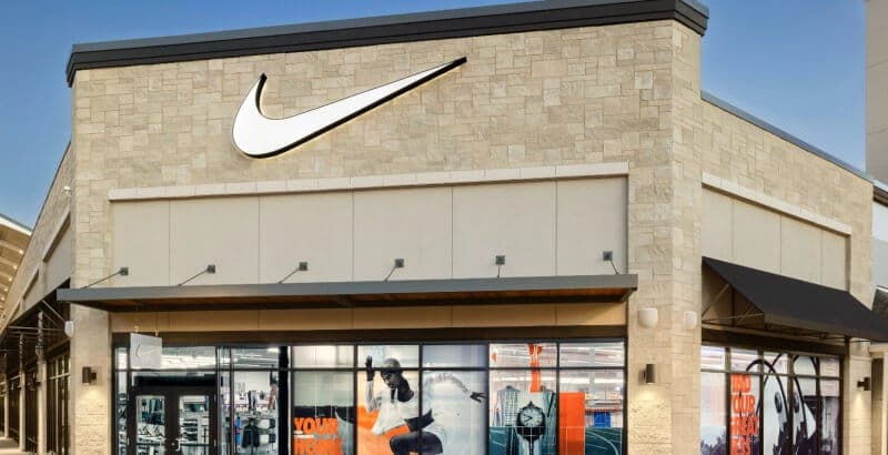 The image shows a Nike store located in a shopping center. The store has a modern design with a beige brick facade and a black awning. The Nike logo is prominently displayed on the facade. The store's large windows showcase a variety of Nike products, including apparel and shoes. The windows also feature large posters