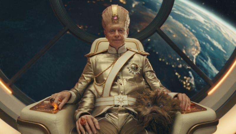 The image shows a man seated in a white, possibly leather, chair with gold accents, in a space setting. He is wearing a gold military-style uniform with a white sash and a gold crown with a pink gem. The man has a light brown or tan skin tone. Next to him, a furry creature resembling a chupacabra or similar animal is s