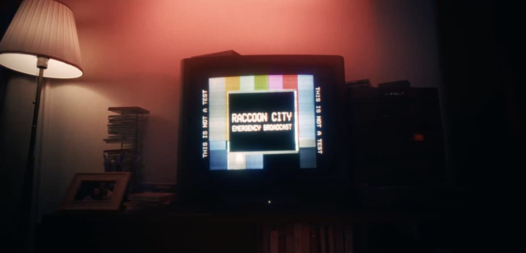 The image shows a television set displaying a message that reads "This is not a test. Raccoon City Emergency Broadcast". The television is situated on a dark wooden or wood-like surface, possibly a desk or table. To the left of the television, a lamp with a white shade casts a warm glow. On the right side of the televi