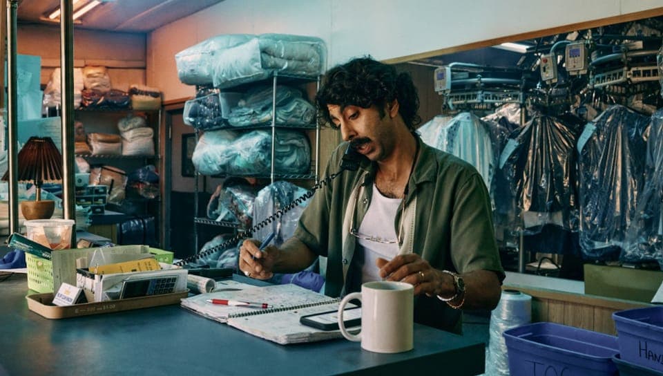 A man with curly hair and a mustache holds a phone receiver and pen at a laundry or dry-cleaning counter, with plastic-wrapped bedding and garments on shelves behind him, a coffee mug, a notebook, and a calculator on the desk. No readable brand text is visible.