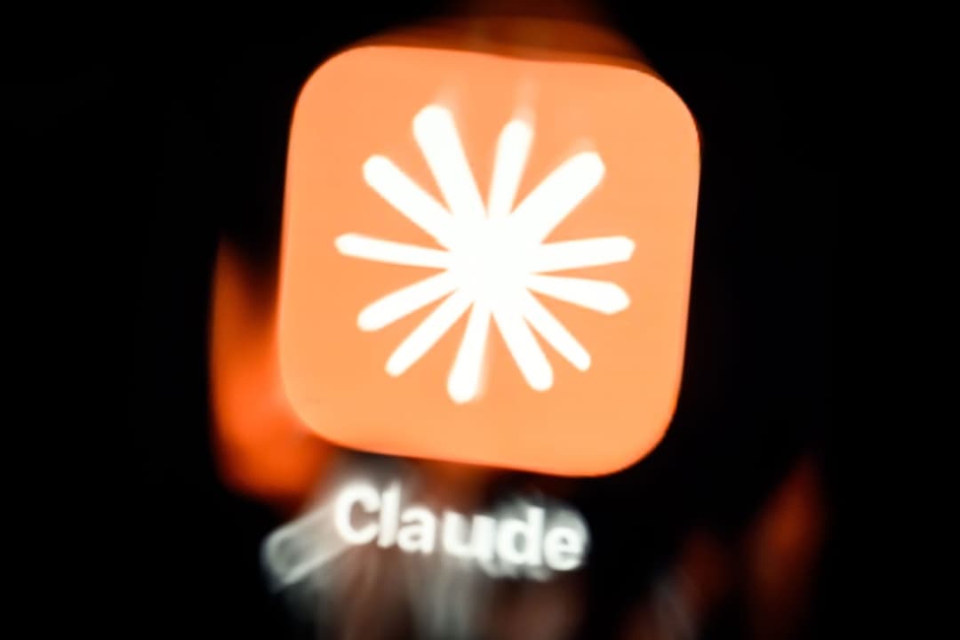 The image shows a close-up view of a person's face, with the word "Claude" written in white text below the face. The face is partially obscured by a bright orange square app icon with a white starburst design. The background is dark, with a blurred orange glow around the app icon, creating a sense of depth and focus on