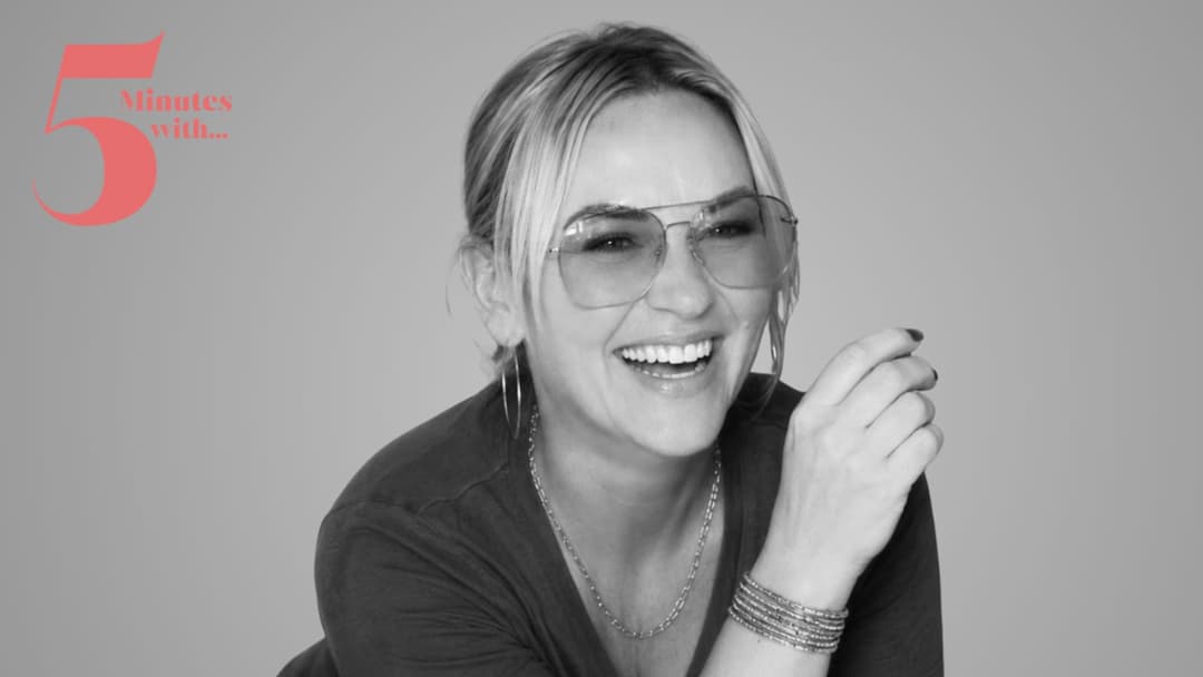The image shows a woman with blonde hair, wearing a dark-colored, possibly black, top and a silver necklace. She is wearing light-colored, possibly pink or light blue, aviator-style sunglasses. She is smiling broadly, with her left hand raised to her face, and her right hand is resting on her left elbow. She is also we