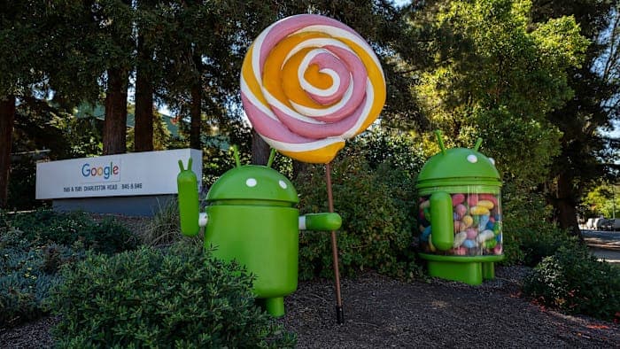 The image shows two large, green Android robot statues positioned in front of a Google sign. The sign, which is white with blue and red text, is located on a grassy area with trees in the background. One of the robots is holding a large, colorful lollipop with a pink and orange swirl design. The other robot is holding