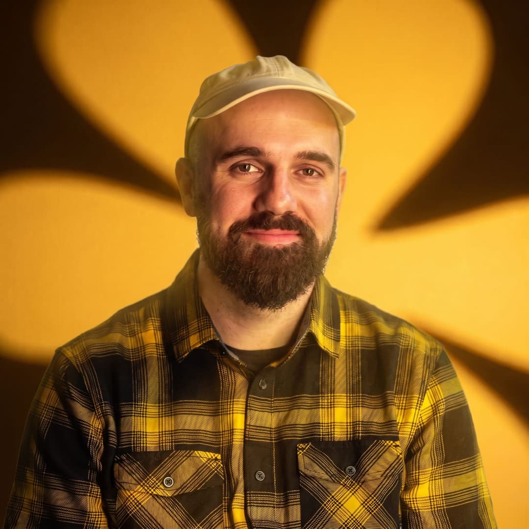 The image shows a man with a beard and a light beige or cream-colored cap, wearing a black and yellow plaid shirt. He is looking directly at the camera with a slight smile. The background is a vibrant yellow, with a large, stylized flower-like shape casting a shadow on the man's face. The man's skin tone is light brown