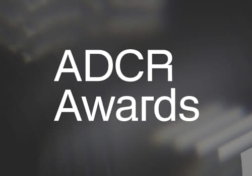 The image presents a stark contrast between the white text "ADCR Awards" and the dark gray background. The text, centrally positioned, is the only element in the image, standing out against the monochrome backdrop. The background is a dark gray, with a blurred, out-of-focus effect, creating a sense of depth and dimensi