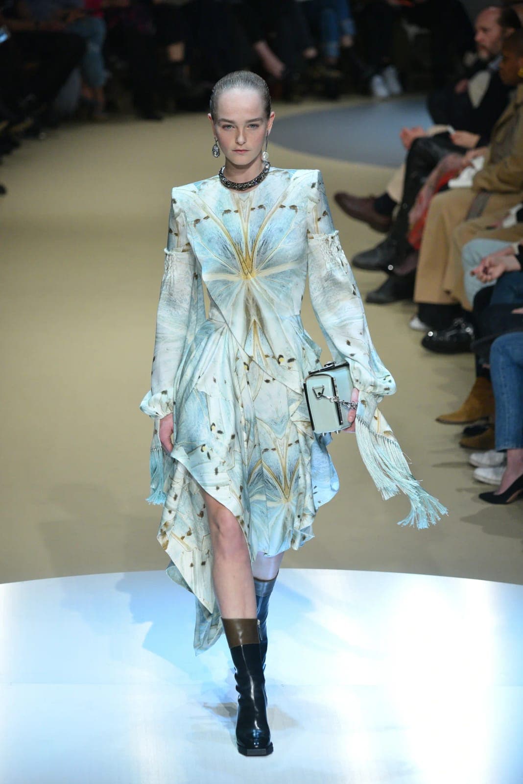 The image captures a model striding down a runway during a fashion show. The model is wearing a light blue dress with a butterfly-like pattern and a long, flowing skirt. The dress features a high neckline and long sleeves, and the skirt has a ruffled hem. The model is also wearing black knee-high boots and a dark-color