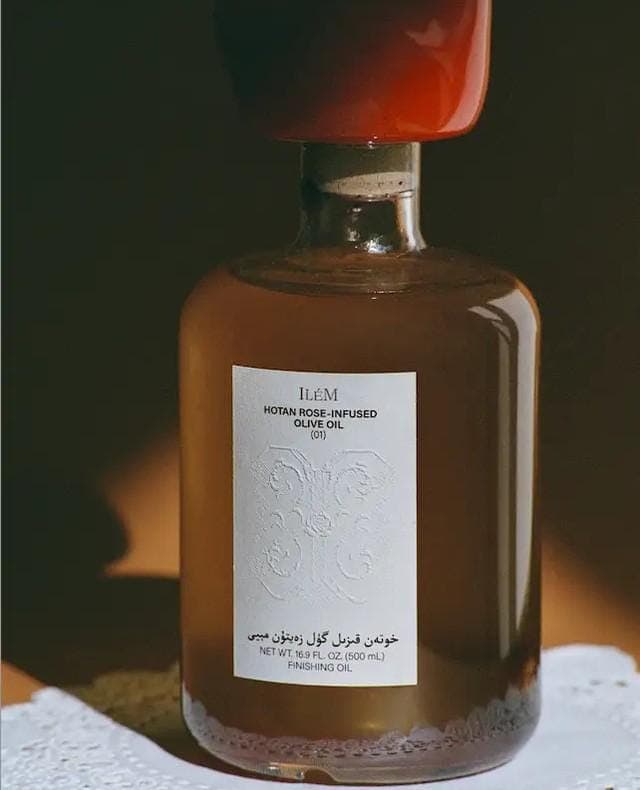 The image shows a bottle of olive oil with a red cap. The bottle is made of glass and has a dark brown or amber liquid inside. The label is white with black text and features a stylized floral design. The label also includes the text "IEM HOTAN ROSE OIL" and "OLIVE OIL (1)". The label also includes the text "����و�� ال