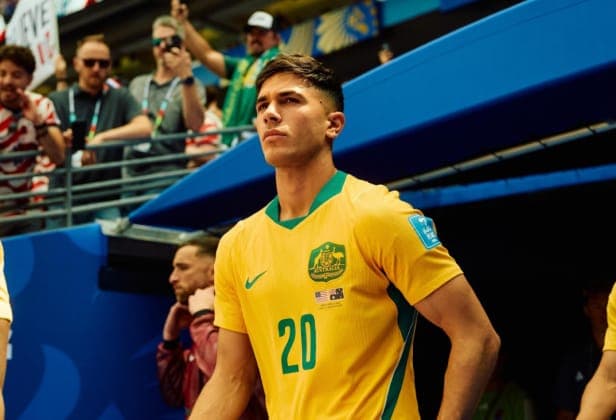 The image shows a young man wearing a vibrant yellow soccer jersey with the number 20, standing in a stadium. The jersey features a green and gold emblem on the chest and a small American flag patch on the sleeve. He has short, dark brown hair and is looking off to the side with a neutral expression. The stadium is fil