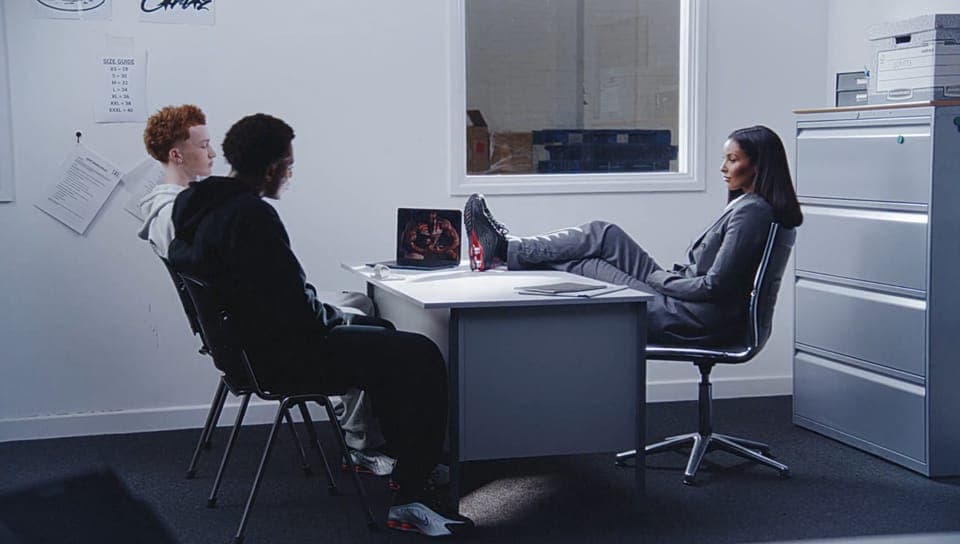 The image shows a group of three individuals seated around a light gray desk in a room with white walls. The person on the left is wearing a black hoodie and has their feet propped up on the desk. The person in the middle is wearing a dark gray or black jacket and has their feet resting on the desk. The person on the r