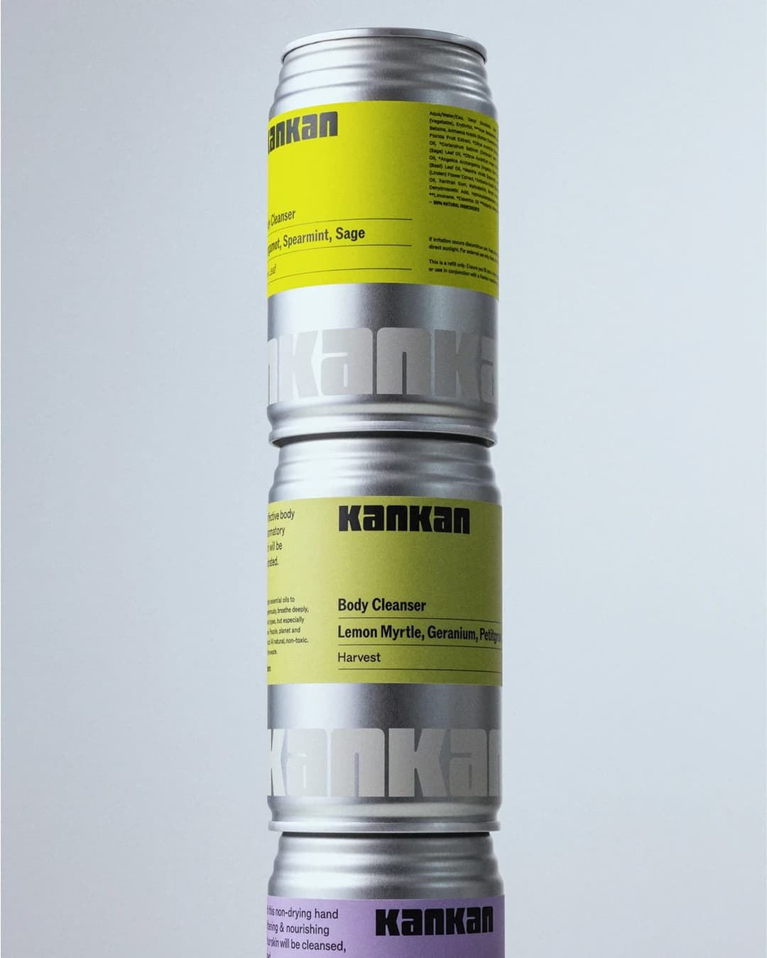The image shows a stack of three Kankan canisters, each with a distinct color and label. The topmost canister is yellow and features the text "Kankan Cleanser" and "Lemon Myrtle, Geranium, Petitgrain". The middle canister is silver and displays the text "Kankan Body Cleanser" and "Lemon Myrtle, Geranium, Petitgrain". T
