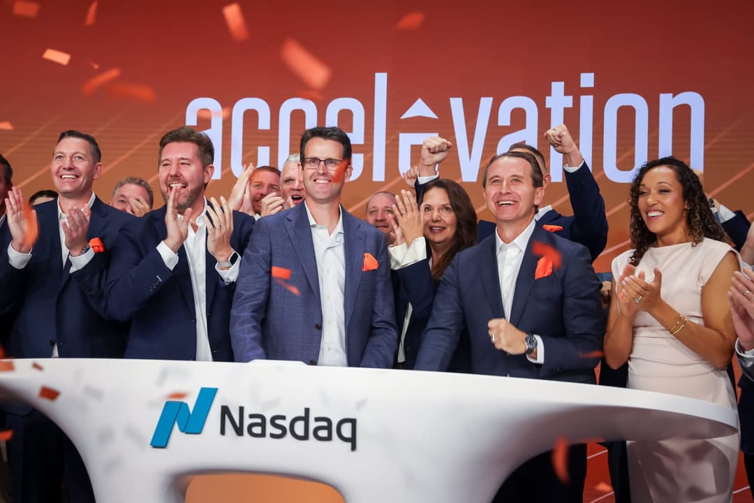The image captures a group of people, including two men in suits and one woman in a light-colored dress, standing behind a white podium adorned with the blue and white logo of the Nasdaq. The podium is positioned in front of a large screen displaying the words "acceleration" in white letters against a red background. T