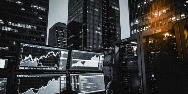 The image depicts a modern office building with a dark, sleek design, featuring multiple tall glass and steel skyscrapers. The building is illuminated by numerous windows, casting a soft glow on the surrounding area. In the foreground, a server room is visible, with a server rack and a network of cables and wires. A pe