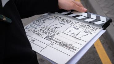 The image shows a person holding a black and white film clapperboard, which is a common tool used in film production. The clapperboard is held in the person's right hand, and the person is wearing a dark-colored, possibly black, suit. The clapperboard is covered in handwritten text in a language that appears to be Russ