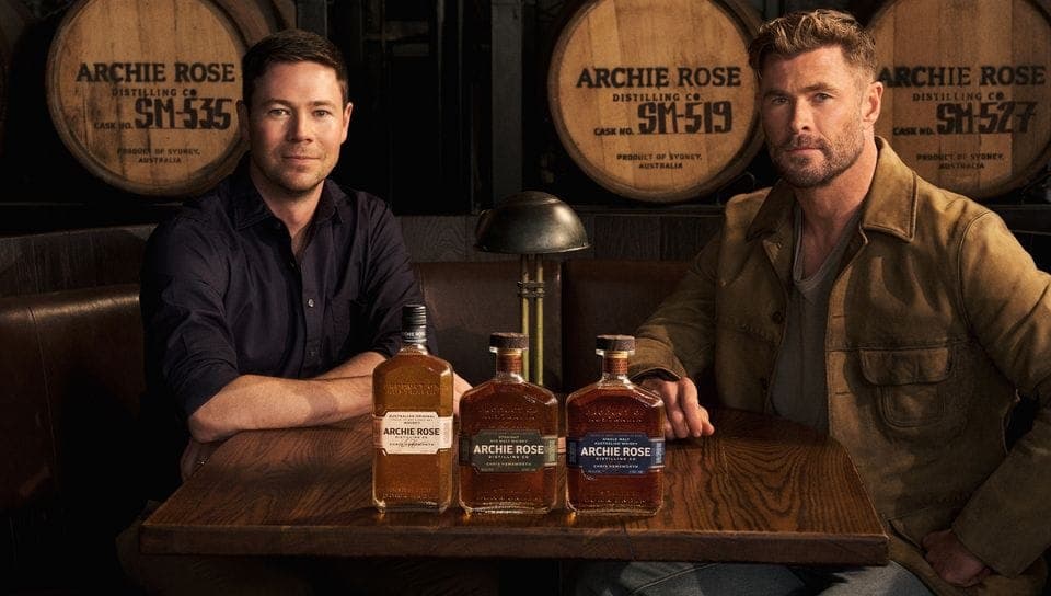 The image shows two men seated at a wooden table in a dimly lit setting, possibly a bar or restaurant. The man on the left is wearing a dark blue or black shirt, while the man on the right is wearing a brown jacket. The table is set with three bottles of Archie Rose Distilling Co. whiskey, each with a unique label. The