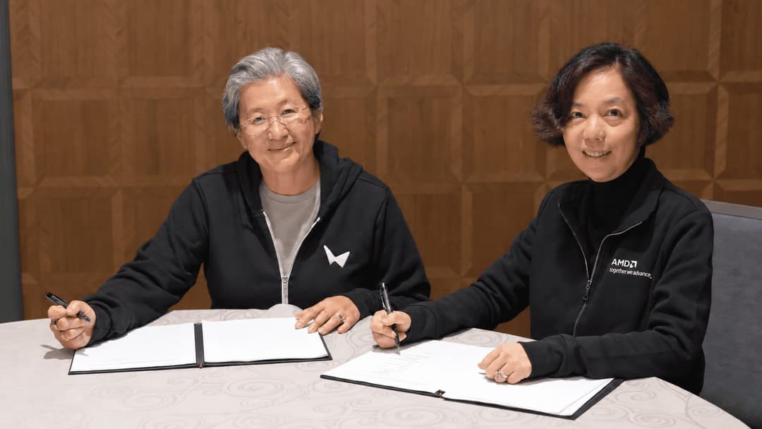The image shows two women seated at a round table, engaged in signing a document. The woman on the left is wearing a black hoodie with a white logo, and the woman on the right is wearing a black jacket with "AMCD" and "Together we advance" written on it. Both women are holding pens and appear to be focused on the docum