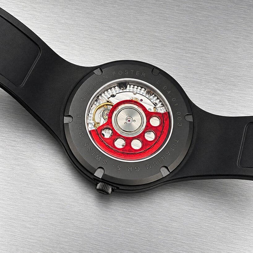 The exposed caseback of a black titanium watch shows a red-accented automatic movement and an engraved "NORMAN FOSTER" with "TITANIUM GR 5 · SWISS MADE" and "L.04.01.003" around the edge. The strap is partly visible on a brushed-metal surface.