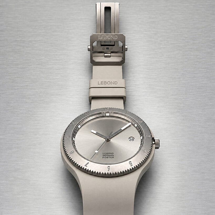 A grey Lebond Norman Foster watch with a titanium-look bezel and a branded "LEBOND" buckle is laid flat on brushed steel, with the strap extended upward. The dial reads "LEBOND NORMAN FOSTER" and the date window shows 28.