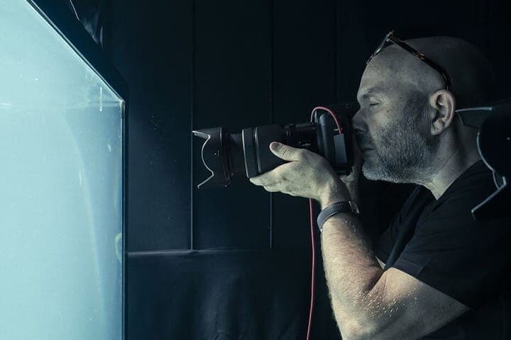 The image shows a bald man with a beard and glasses, wearing a black t-shirt, holding a camera with a red lens and a black body. He is positioned in front of a dark, possibly black, wall or backdrop, with a bright blue screen or window to his left. The man is looking intently at the screen, which appears to be a photog