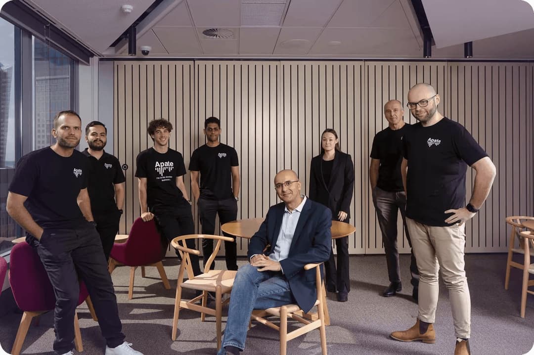 A group portrait of eight people posed in a modern office with a wooden slatted wall, wooden Wishbone-style chairs, and red upholstered chairs; most wear black T-shirts with the white "Apate" logo, and one shirt reads "I'm in the decline business...".