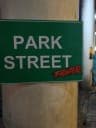 The image shows a green street sign with white lettering that reads "PARK STREET" and features a red "FIGHTERS" logo. The sign is attached to a light brown or beige pole. The background is out of focus, but it appears to be an industrial or urban setting.