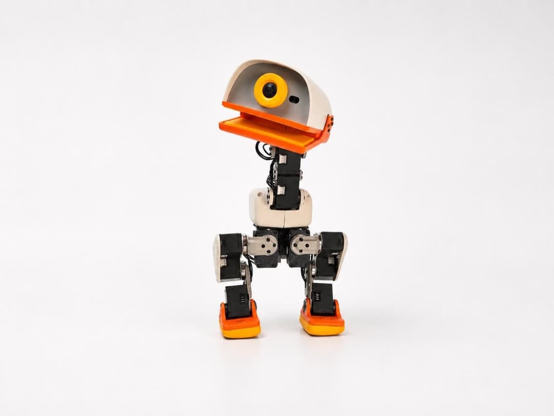 The image shows a small robot standing upright on a white background. The robot has a primarily white body with black and orange accents, including a large orange beak and orange feet. It has a yellow circular head with a black dot, and a black body with orange accents. The robot's arms are black and extend outwards, a