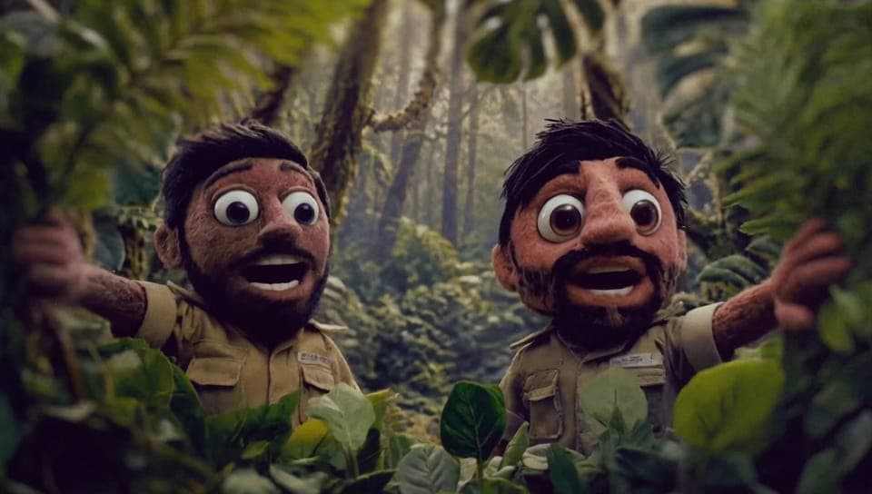 Two felt puppet characters with large googly eyes and beards, wearing khaki safari-style shirts, stand in a dense jungle with leaves in the foreground. No visible text or brand.