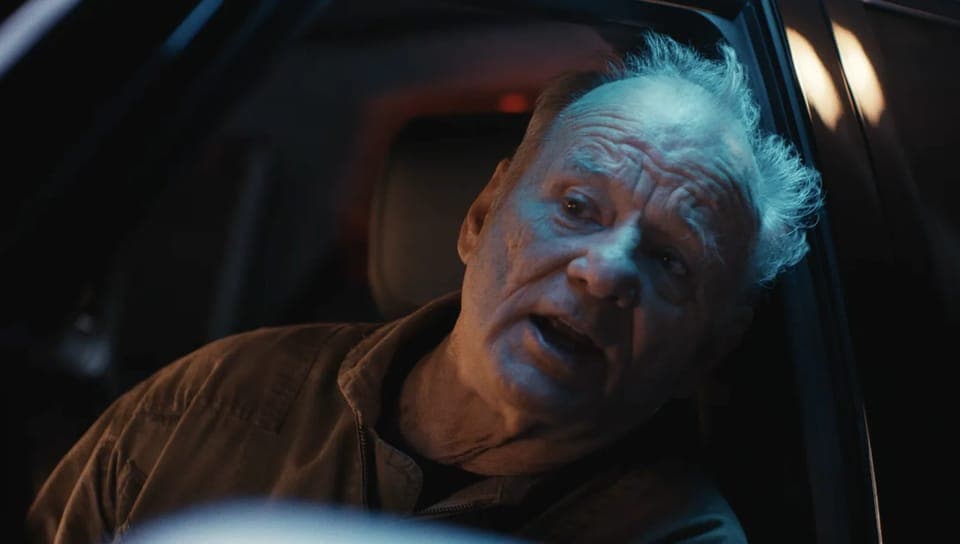 The image shows an older man with a weathered face, wearing a brown jacket, seated in the driver's seat of a car. His hair is a light gray or white color, and his eyes are wide open, giving the impression of surprise or alarm. The car's interior is dark, with the backseat and headrests appearing black. The scene is ill