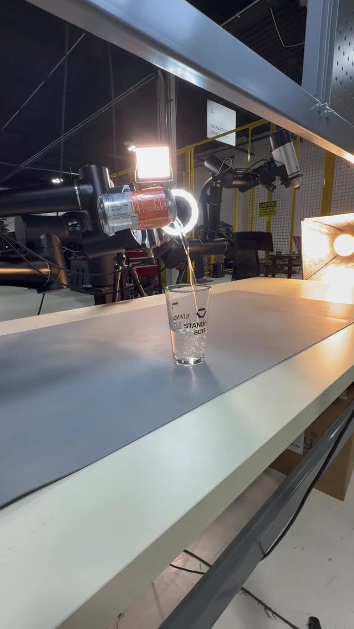 The image shows a robotic arm pouring a clear liquid from a can into a clear glass on a white table in a factory setting. The table is situated on a gray conveyor belt, and the background reveals a workspace with various industrial equipment, including a yellow caution sign and a yellow light fixture. The robotic arm i
