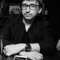 A black-and-white portrait of a man with dark-rimmed glasses and a dark jacket, seated at a table with a large wristwatch visible on his wrist. No legible text or brand is visible.