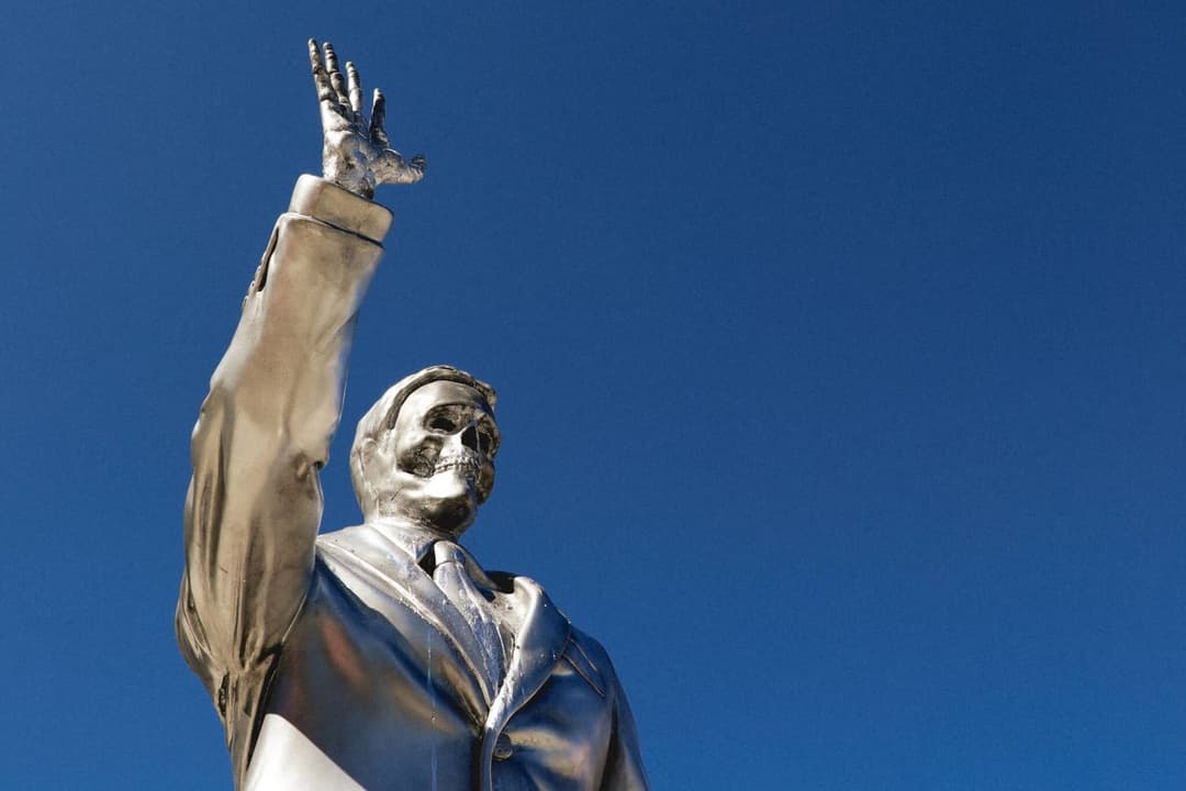 The image depicts a striking silver statue of a man standing against a clear blue sky. The man is wearing a suit and tie, and his right hand is raised in a gesture of victory or triumph. The statue is positioned slightly off-center in the frame, with the man's head and upper body dominating the composition. The statue