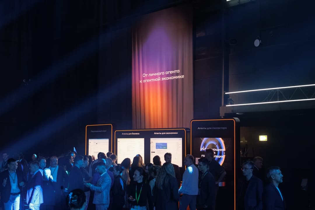 The image captures a bustling scene at a technology conference, with a large group of people gathered around a series of digital displays. The displays, which are primarily white with orange/red accents, are arranged in a row and feature text in a language other than English. The text, which appears to be Russian, is d