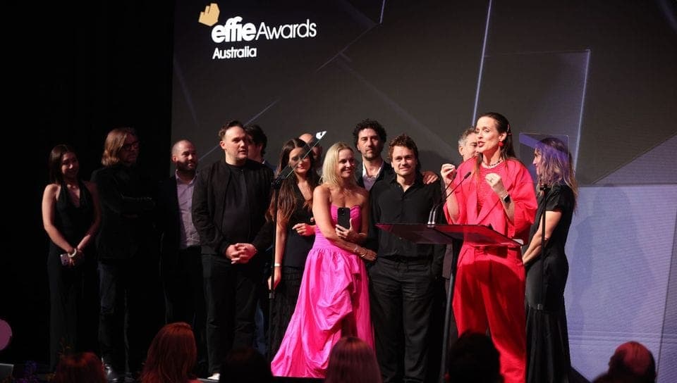 The image captures a group of people on a stage, with a large screen displaying the text "effie Awards Australia". The stage is set against a dark background with a geometric pattern. The group consists of at least nine individuals, with some standing and others seated. The central figure is a woman in a vibrant pink d