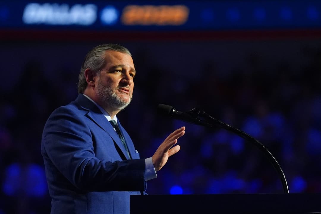 The image shows a man in a blue suit and tie standing at a podium, speaking into a microphone. He has a beard and is gesturing with his right hand. The podium is positioned in front of a large screen displaying a blurred image of a crowd, suggesting a public speaking event. The man's suit appears to be a dark blue or n