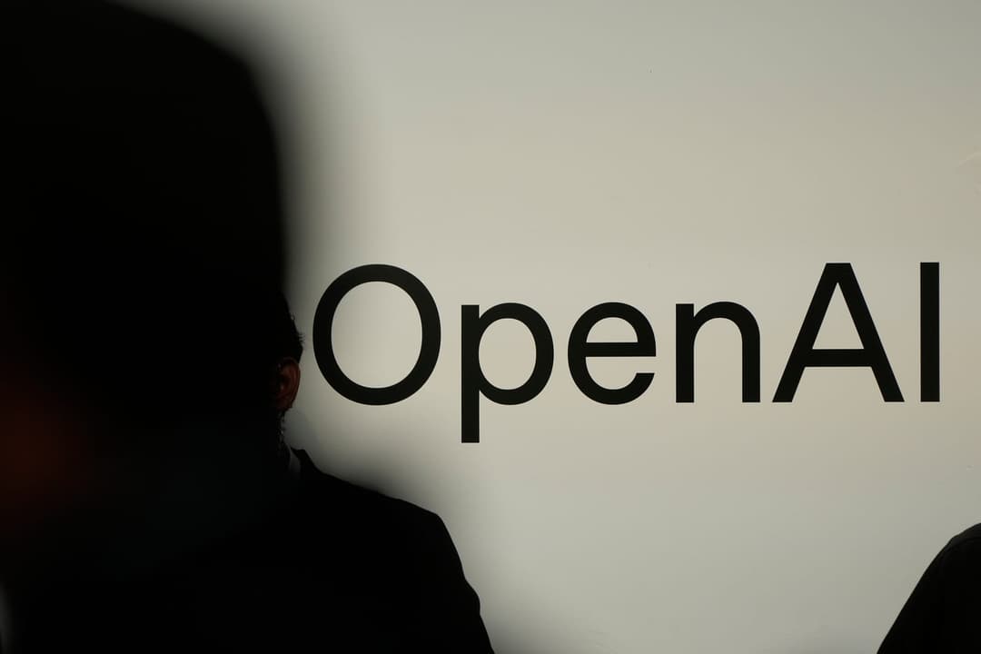 The image shows a person standing in front of a white wall with the words "OpenAI" written in black letters. The person's back is turned to the camera, and their silhouette is visible against the stark white background. The image is taken from a low angle, emphasizing the height of the person and the wall. The lighting