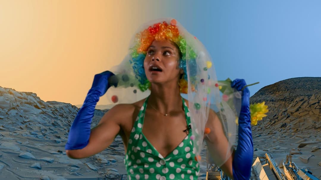 The image shows a woman standing in a desert-like environment, wearing a vibrant clown wig, a green polka-dot dress, and blue gloves. She is holding a small bouquet of yellow flowers in her left hand. The background features a rocky landscape with a large, dark-colored object that resembles a bulldozer or excavator. Th