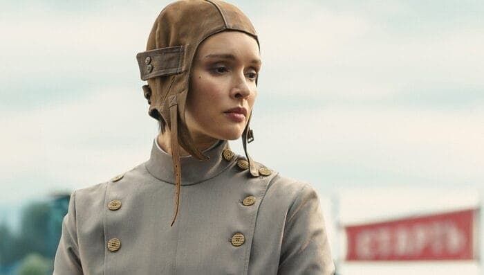The image shows a woman standing in front of a blurred background. She is wearing a light gray or beige jacket with gold buttons and a brown leather hat with a chin strap. The hat has a dark brown or black band around the base of the chin strap. The woman's gaze is directed off to the side, and she has a neutral expres