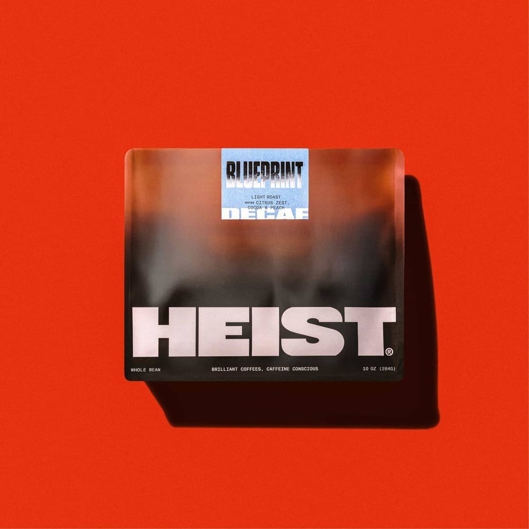 The image shows a single coffee bean bag bag resting on a vibrant red background. The bag is primarily black with a gradient of orange and brown tones, creating a visually striking contrast. The word "HEIST" is prominently displayed in bold white letters on the bag. The bag also features a small, light blue label with