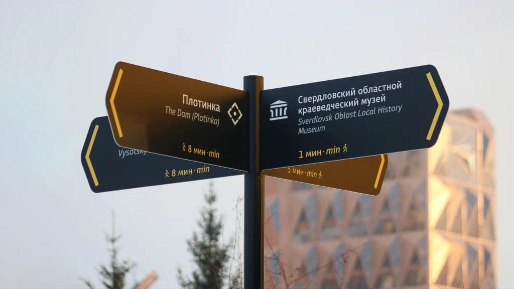 The image shows a black and gold directional signpost standing against a clear blue sky. The signpost has three arms, each pointing in a different direction. The left arm points towards "The Dam (Plotkinja)" and the right arm points towards "Sverdlovsk Oblast Local History Museum". The middle arm points towards "������