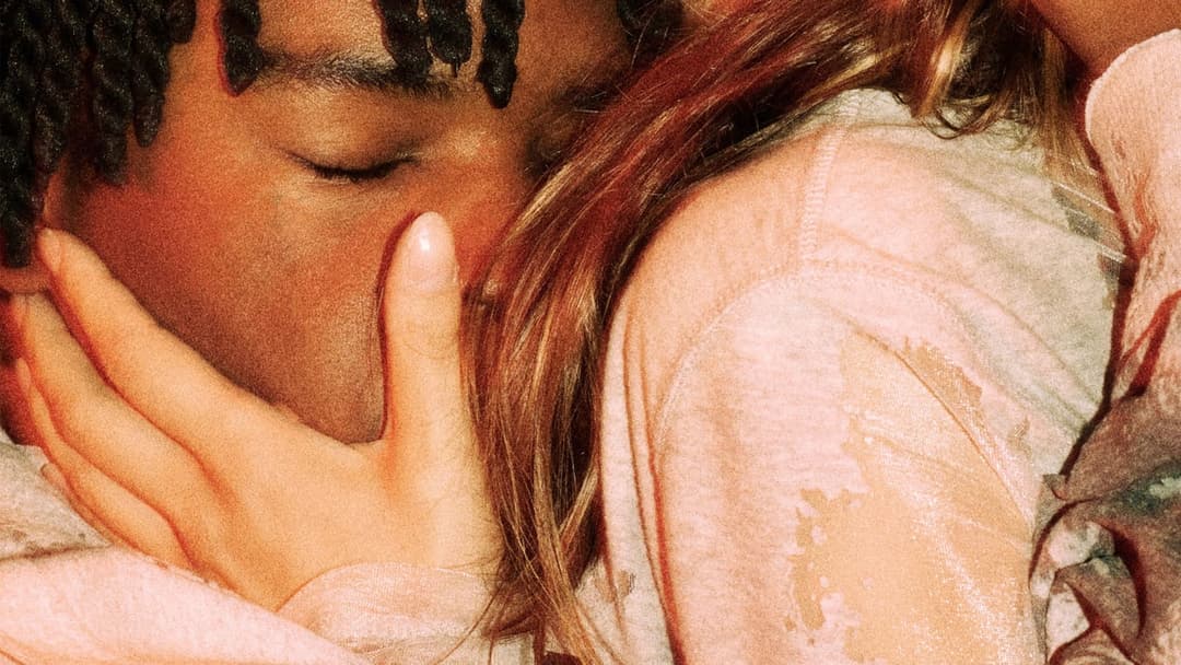 The image shows a close-up of a person's face, with their eyes gently closed and their head resting on their hand. The person has dark brown skin and dark, braided hair. The hand is positioned over the person's shoulder, and the person is wearing a light pink or peach-colored shirt. The person's arm is draped over the