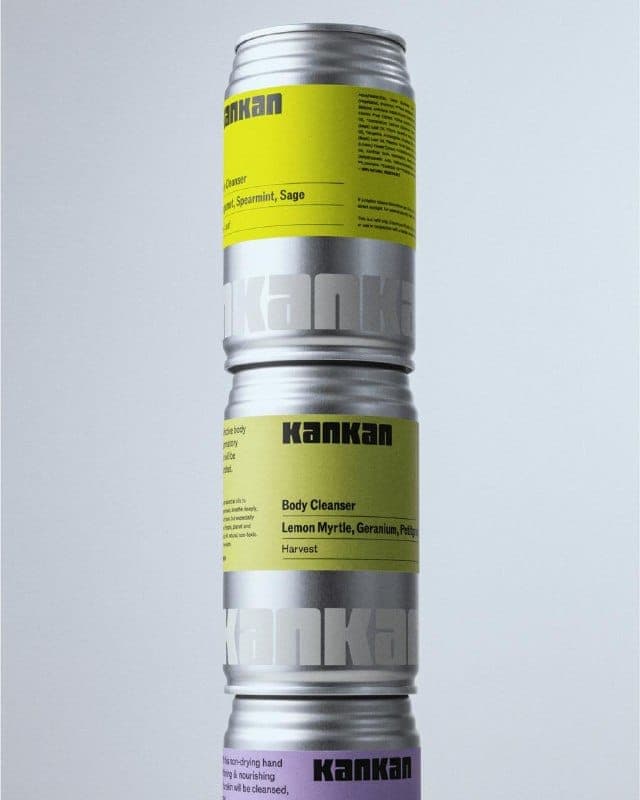 The image shows a stack of three Kankan products, each in a silver can. The top can is yellow and labeled "Body Cleanser Lemon Myrtle, Geranium, Petitgrain, Harvest". The middle can is light green and labeled "Body Cleanser Lemon Myrtle, Geranium, Petitgrain, Harvest". The bottom can is light purple and labeled "No-dry